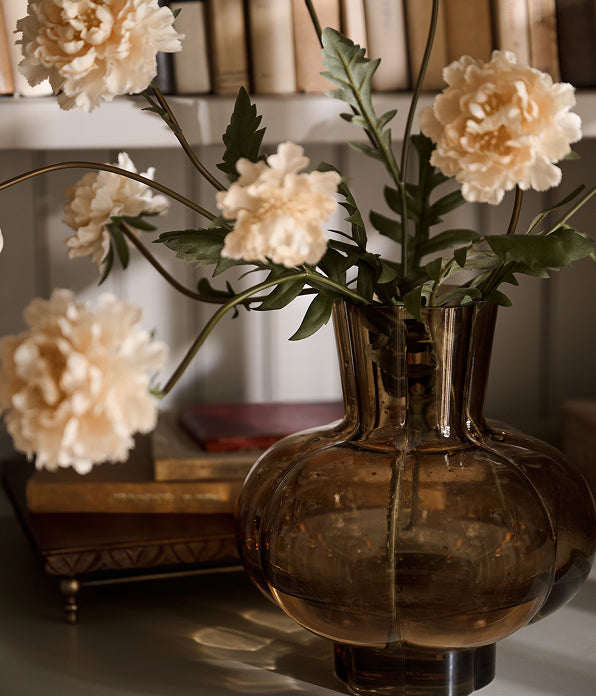 A dark amber glass vase filled with cream-colored artificial peony flowers and green stems, styled on a shelf in front of a blurred background of books.
