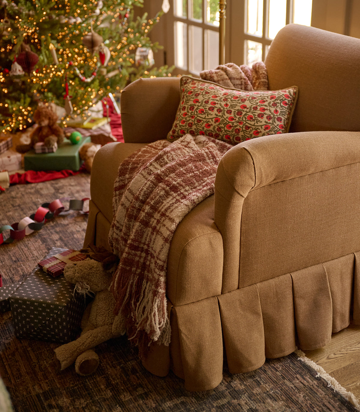 A beige upholstered armchair in a cozy holiday living room, styled with a red and brown plaid throw blanket, a patterned accent pillow, and a skirted base. A decorated Christmas tree, wrapped gifts, and a teddy bear are visible in the background.