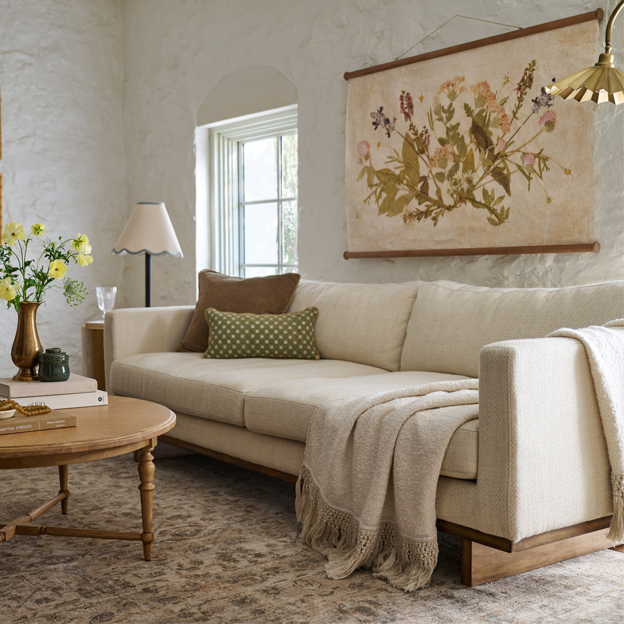 ivory sofa with two throw pillows and an ivory throw laying across.  hanging above it is a pressed floral wall tapestry.