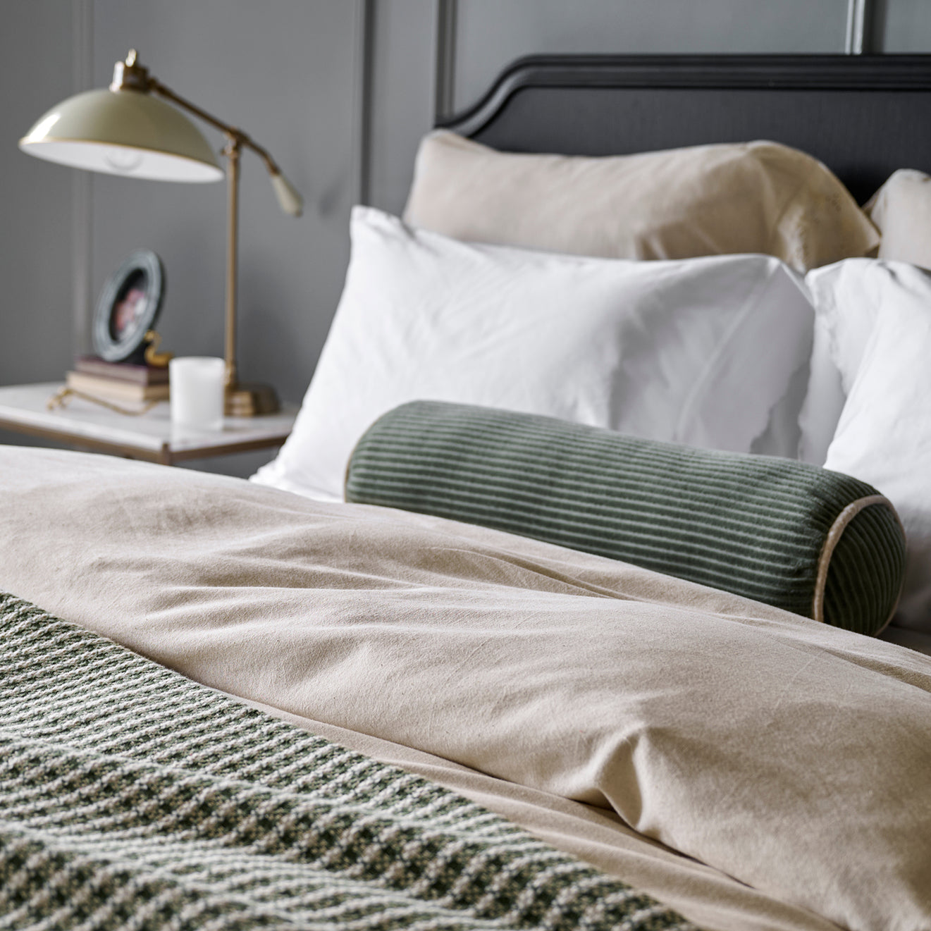 Neatly made bed with white pillows and a green cylindrical pillow, nightstand with lamp and photo frame in the background.