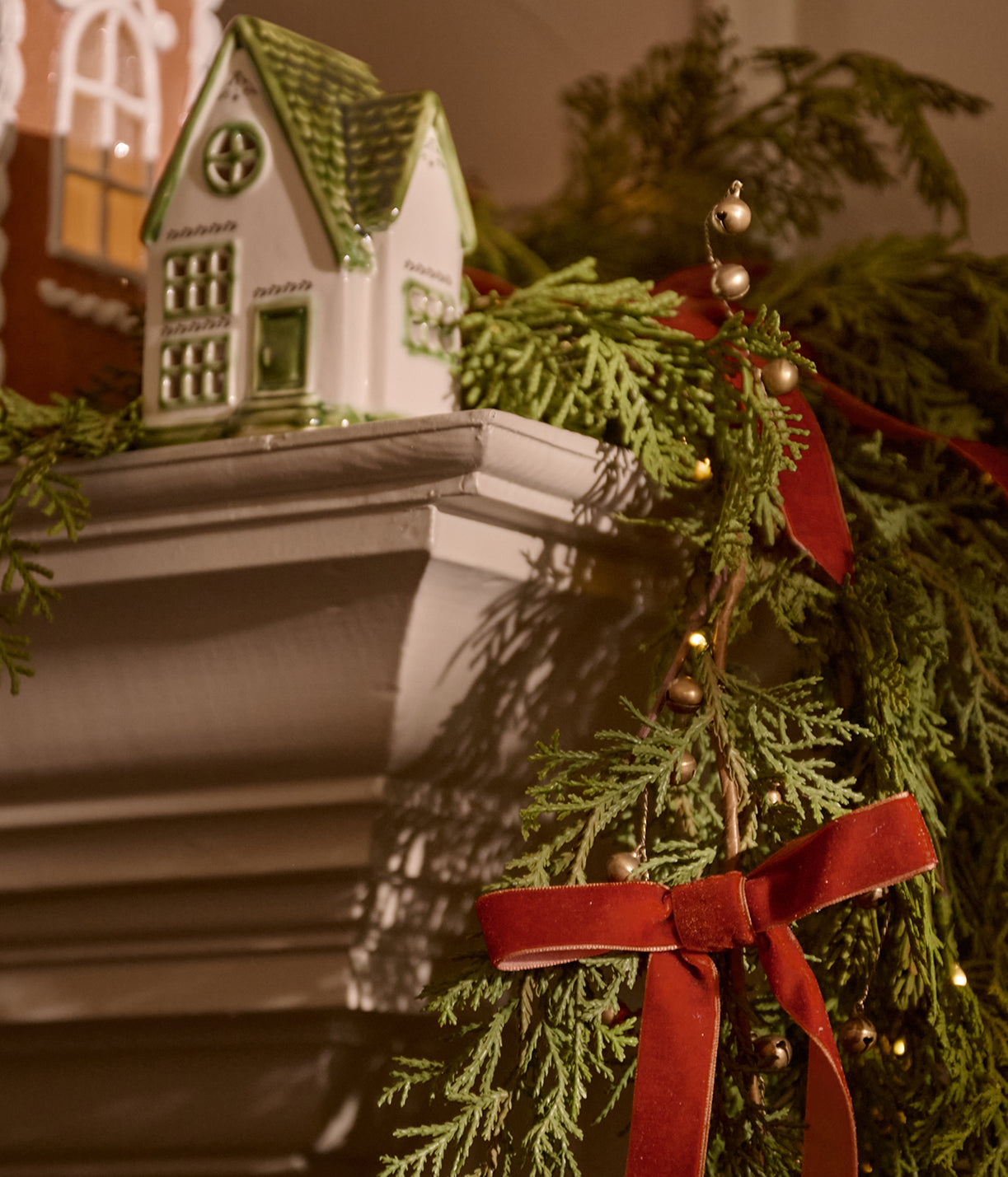 A close-up festive scene showing a white ceramic gingerbread house ornament resting on a white mantel, surrounded by green evergreen garland, red velvet ribbons, and small gold ornaments.