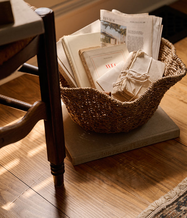 A woven natural-fiber basket filled with folded papers, a magazine, and a small tied bundle, placed on a stone slab on a wooden floor beside a chair leg.