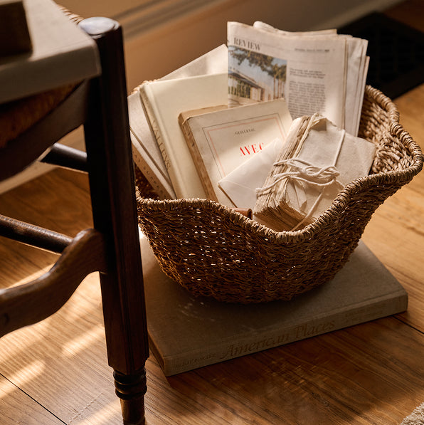 A woven natural-fiber basket filled with folded newspapers and small wrapped paper bundles, placed on a large book on a wooden floor in a warm indoor setting.
