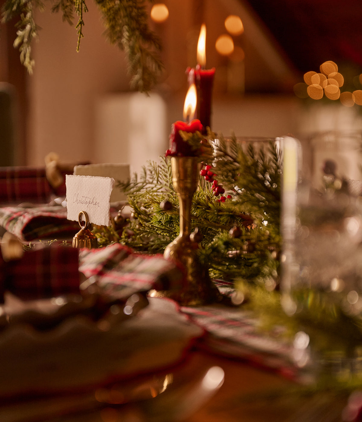 A warm, festive tabletop setting with green pine garlands, lit red candles in brass holders, plaid napkins, and glassware in a softly blurred indoor environment.