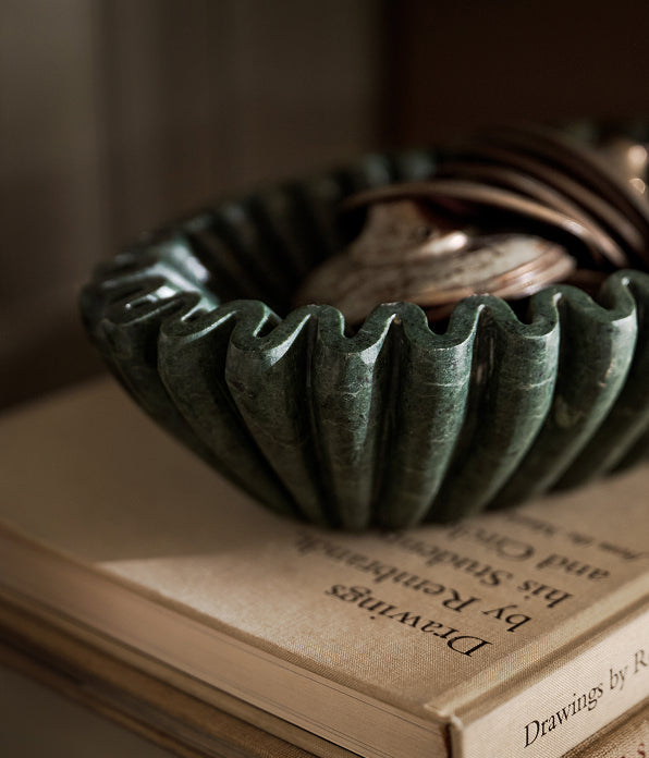 A dark green fluted stone bowl filled with coins, resting on top of an open book in a warm, softly lit setting.