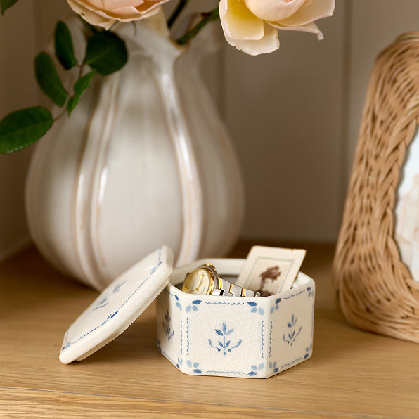 Small decorative box with jewelry on a wooden surface, with a vase and basket in the background.