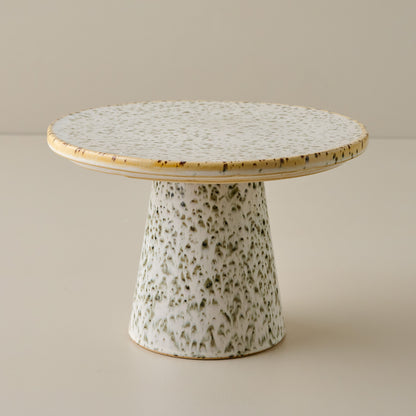 Green speckled ceramic cake stand with a round top and cylindrical base on a beige background