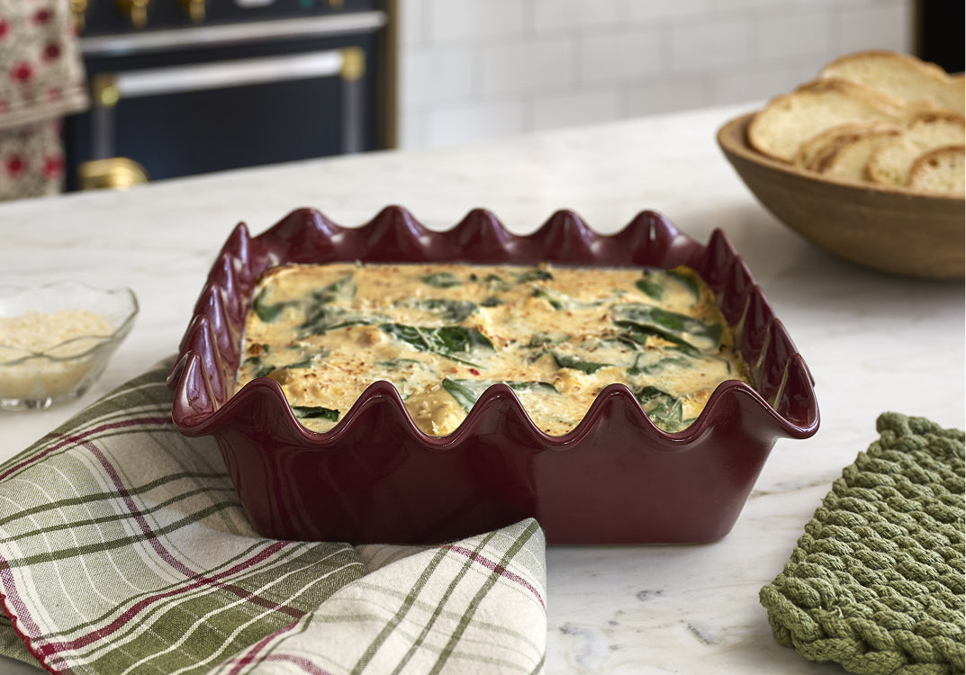 Square scalloped dish of quiche with spinach on a marble countertop, with a bowl of bread in the background.