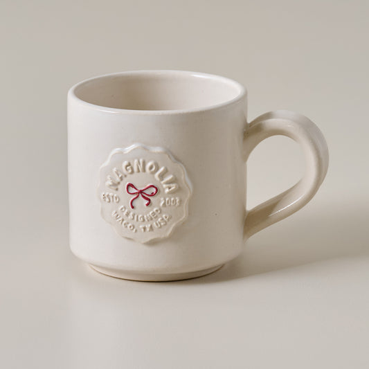 White mug with Magnolia logo and red bow on a light background