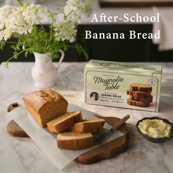 Loaf of banana bread with slices on a wooden board, Magnolia Table packaging, and flowers in the background.