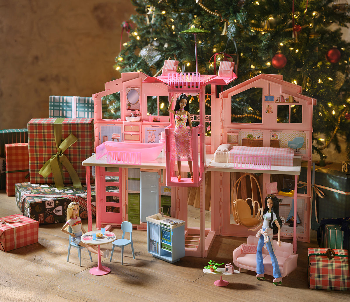 A pink Barbie-style dollhouse with multiple rooms, a balcony, miniature furniture, and the Joanna Gaines Barbie doll, staged in front of a decorated Christmas tree with wrapped gifts on a wooden floor.