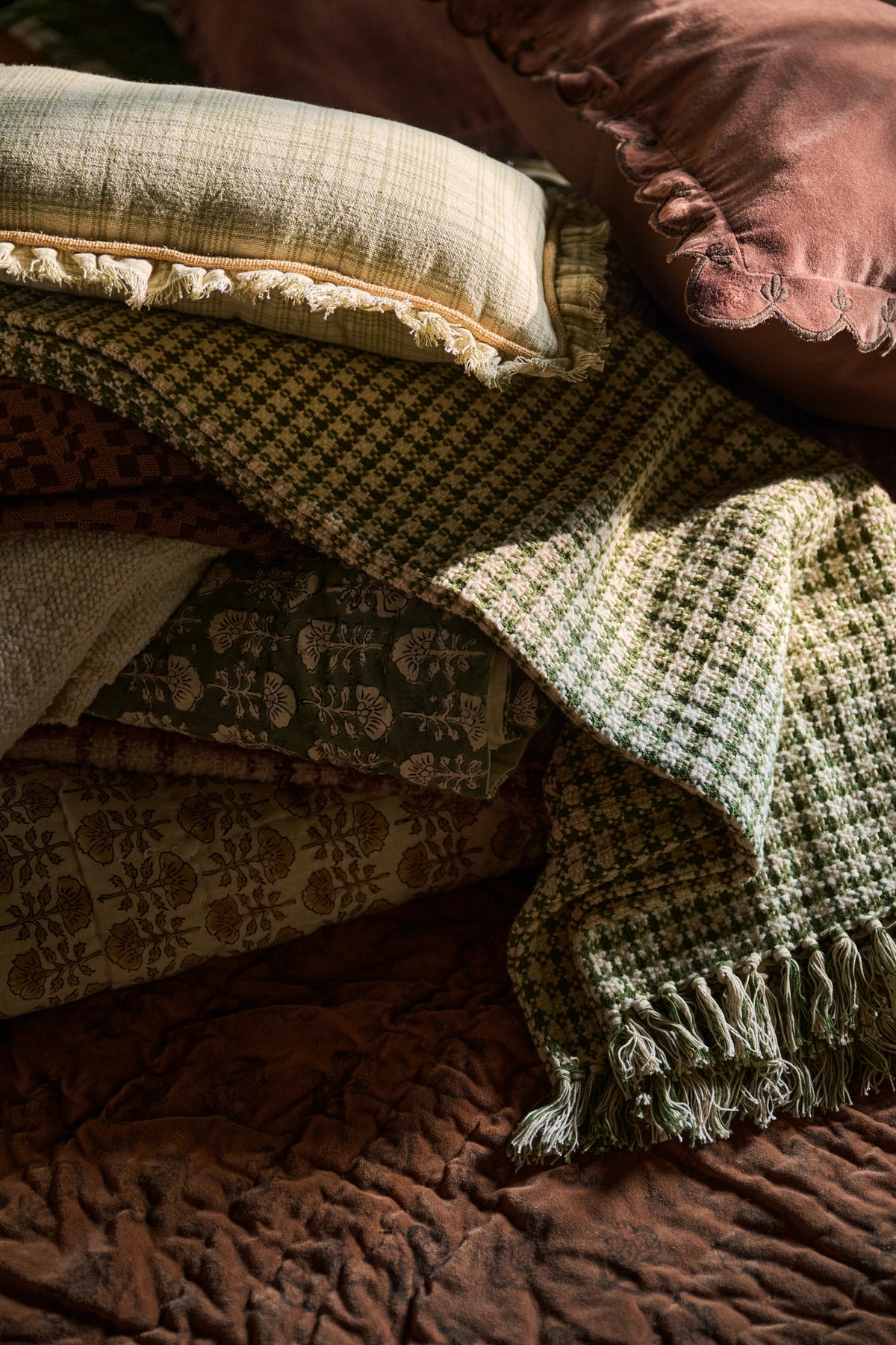 A styled collection of cozy home textiles, including a green and beige houndstooth throw blanket with fringe, a patterned olive green and beige fabric, a cream pillow with fringed edges, and a dusty rose velvet pillow, all arranged on a brown quilted bedspread.