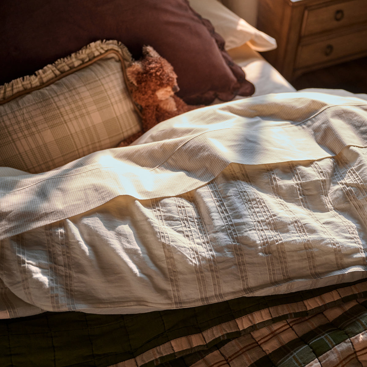A close-up of a quilted cream and beige comforter or duvet on a bed, styled with plaid pillows, a brown pillow, and a small teddy bear in a warm, sunlit bedroom setting.