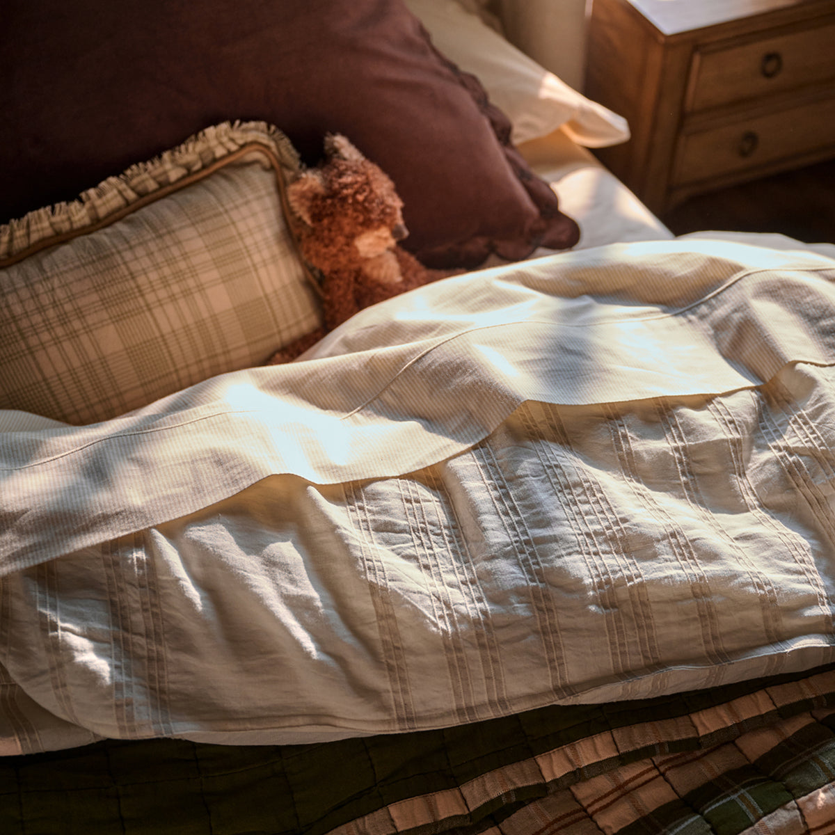 A close-up of a bed with cream-colored textured bedding, including a quilt or comforter and matching pillowcases, styled with brown and beige plaid pillows and a small teddy bear in a warm, sunlit bedroom setting.
