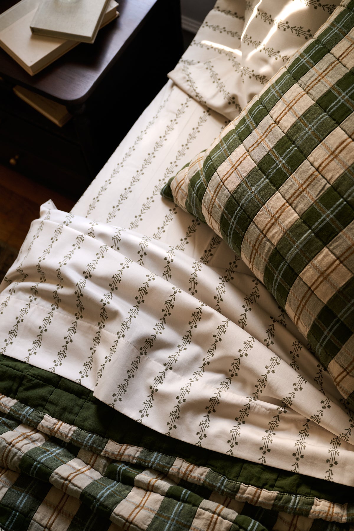 A close-up of a bed styled with a white sheet featuring a delicate green floral vine pattern, a green and beige plaid pillow, and a matching green plaid quilt, with a dark wooden bedside table in the background.