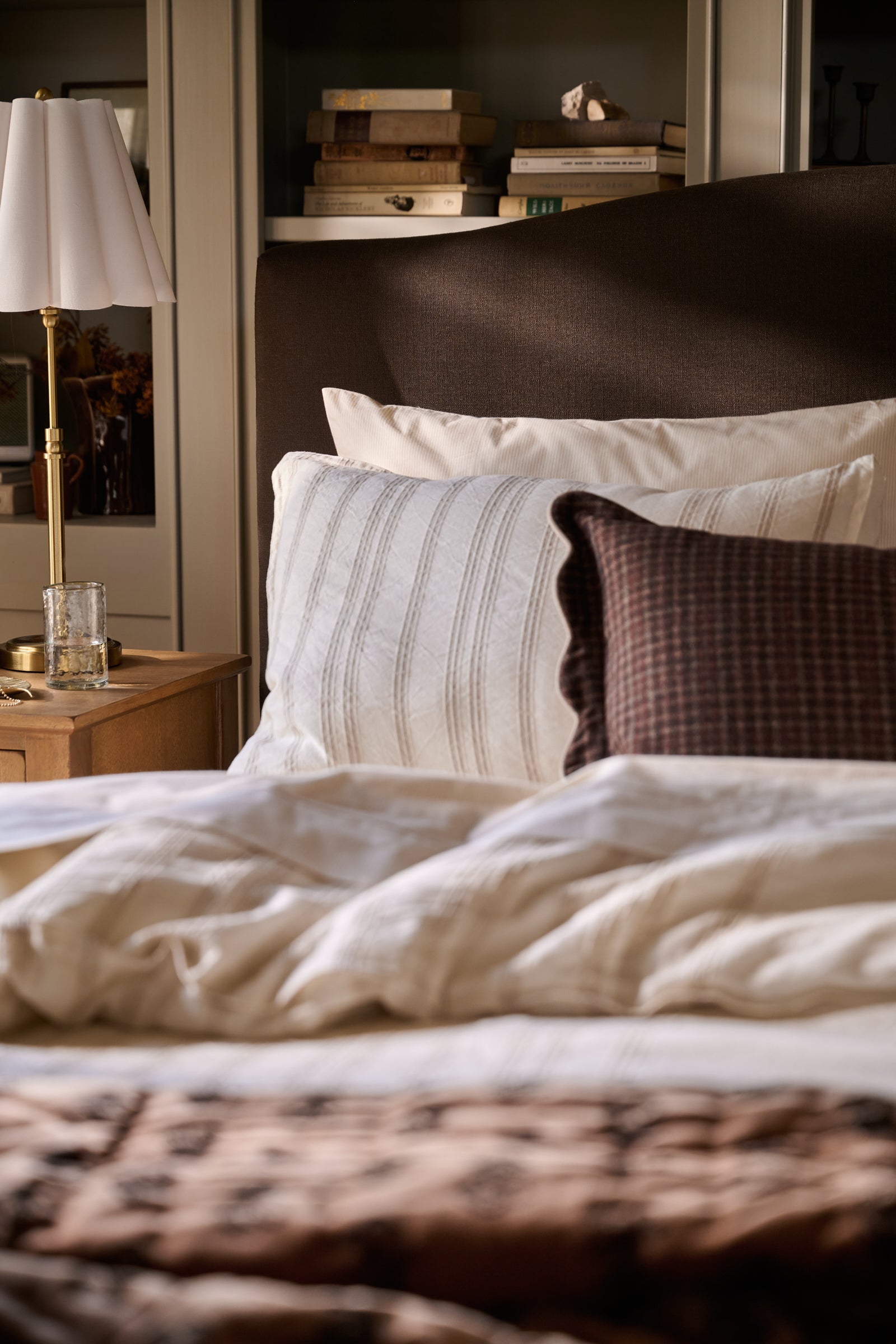A close-up of a bed in a styled bedroom setting, featuring cream and white striped pillows, a dark brown textured headboard, a white bedside lamp on a wooden nightstand, and a soft brown blanket in the foreground.