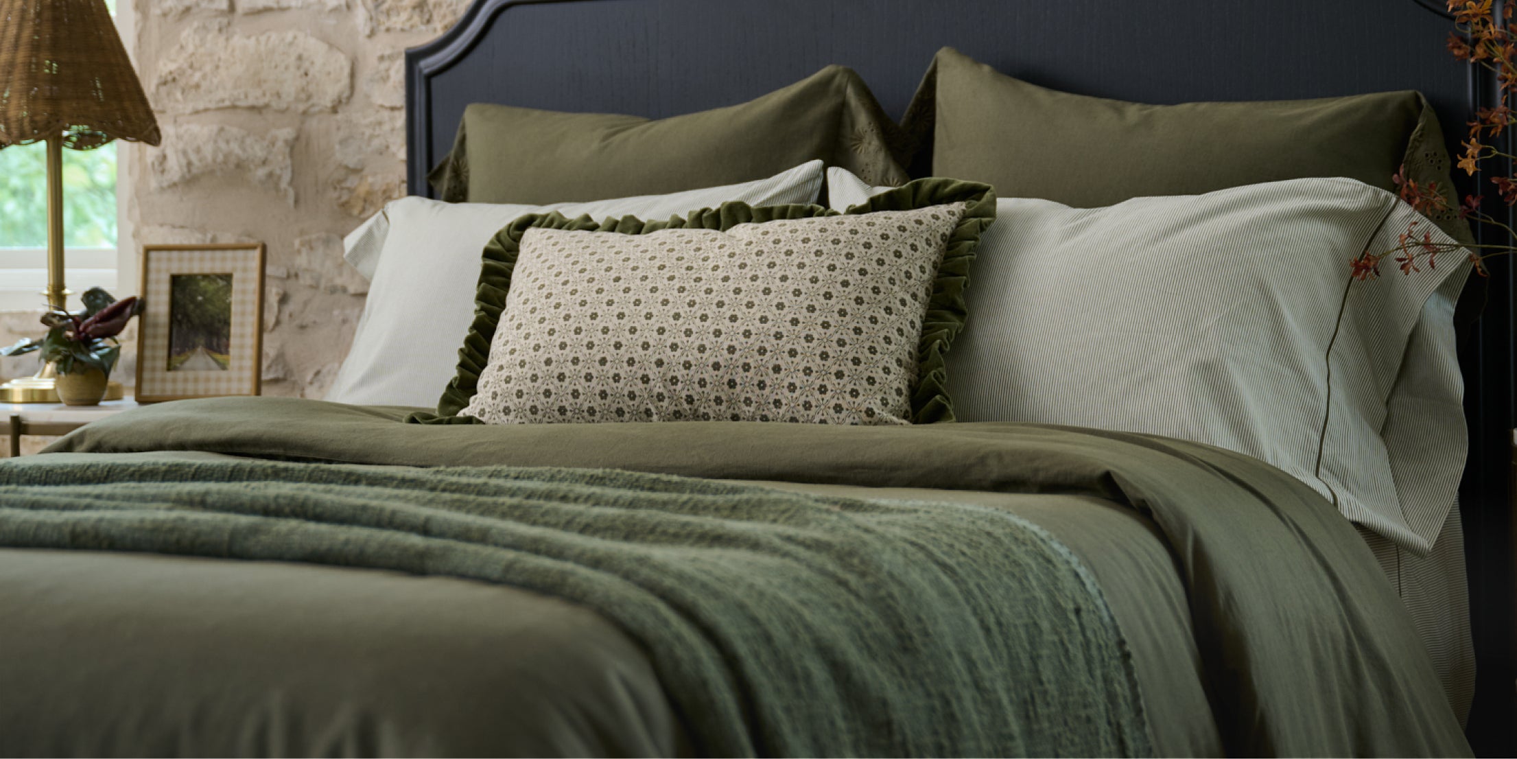 Bed with green bedding and pillows against a stone wall.