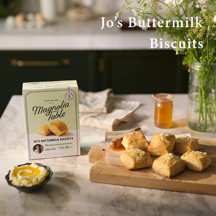Box of Magnolia Table Frozen Jo's Buttermilk Biscuits on a kitchen island with six baked Jo's buttermilk biscuits on a cutting board with honey and butter and flowers. Jo's Buttermilk Biscuits.  Learn More.