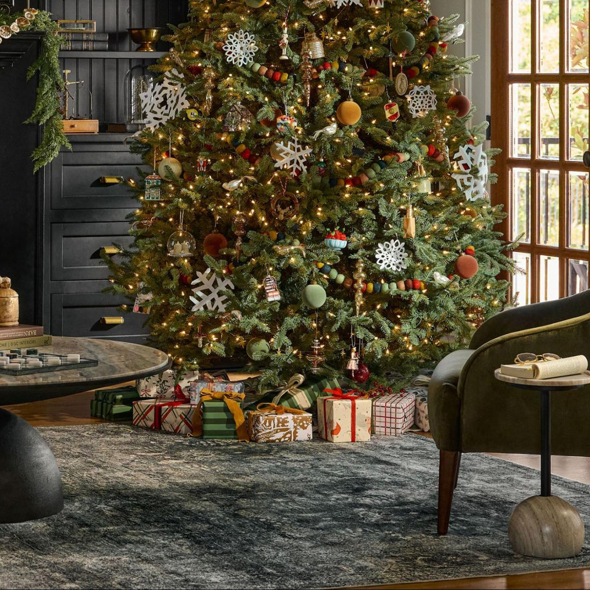A decorated Christmas tree in a cozy living room setting, surrounded by wrapped gifts under the tree and styled with a dark green armchair, a round coffee table, and a patterned area rug.