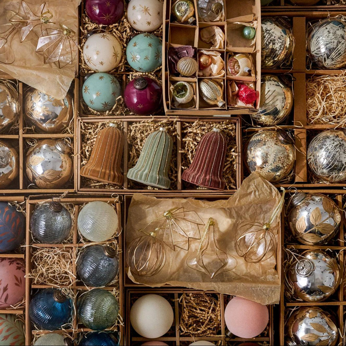 An overhead flat-lay of assorted Christmas ornaments in various colors and textures, displayed inside brown gift boxes with shredded paper. The collection includes metallic silver, blue, pink, green, and gold ornaments, ribbed bells, and hanging glass ornaments.