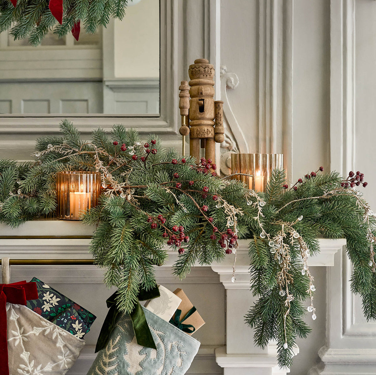 A festive Christmas garland draped across a white fireplace mantel, featuring green pine branches, red berries, lit candles in gold-toned glass holders, and delicate white bead garlands. A wooden nutcracker stands behind the garland, with stockings and wrapped gifts partially visible below.