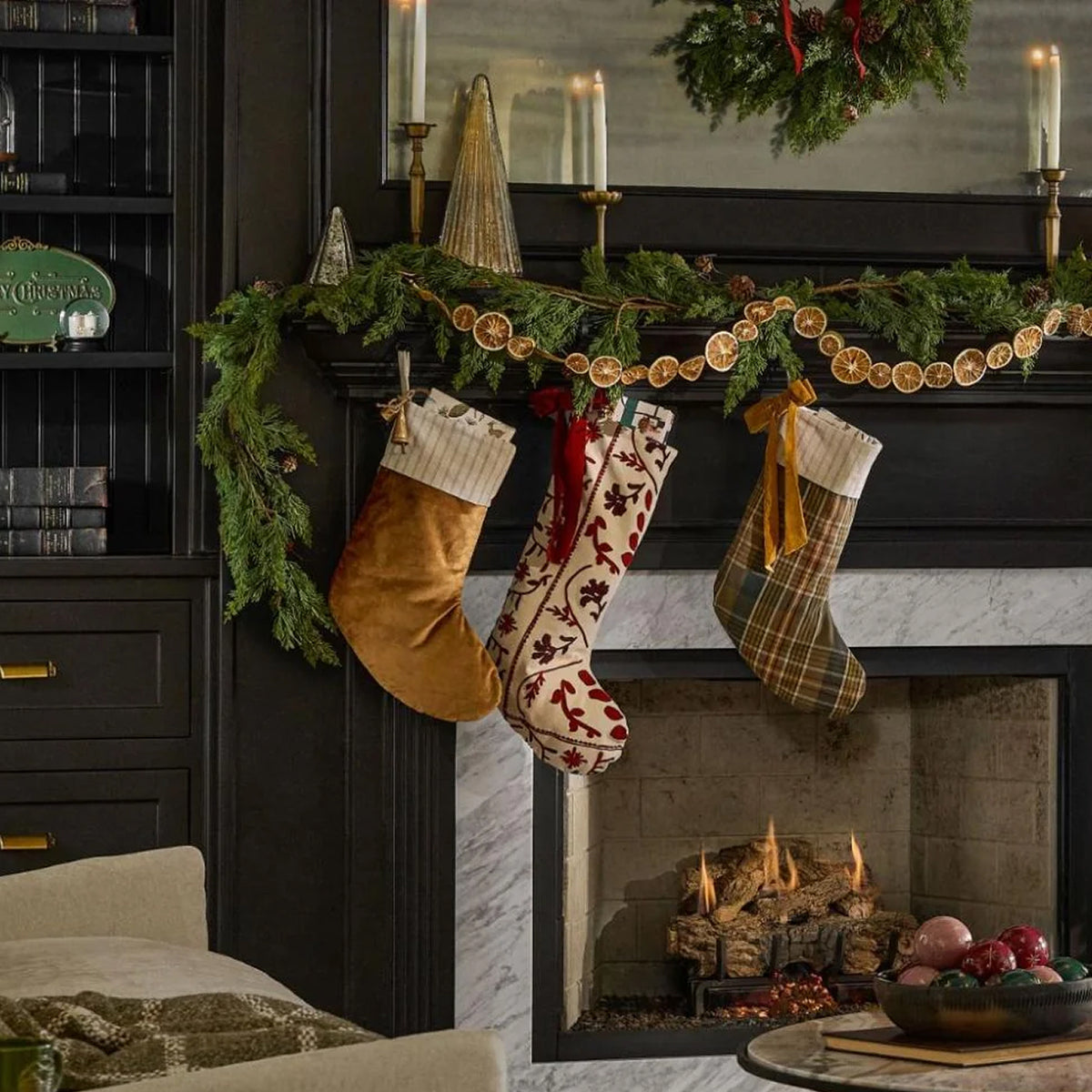 Three festive Christmas stockings hang from a decorated fireplace mantel in a cozy living room. The stockings feature different patterns: one is tan with white stripes, one is cream with red holiday motifs, and one is green plaid.