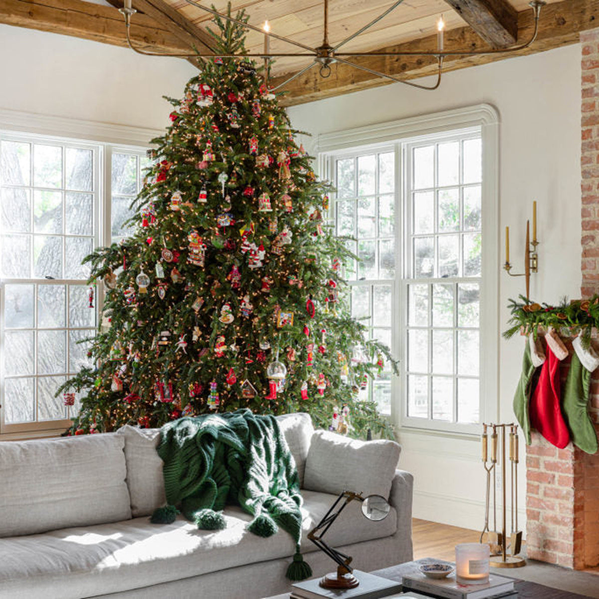 A decorated Christmas tree stands in a bright living room with large windows, a light sofa with a green knit throw, and a brick fireplace with stockings hanging on the mantel.