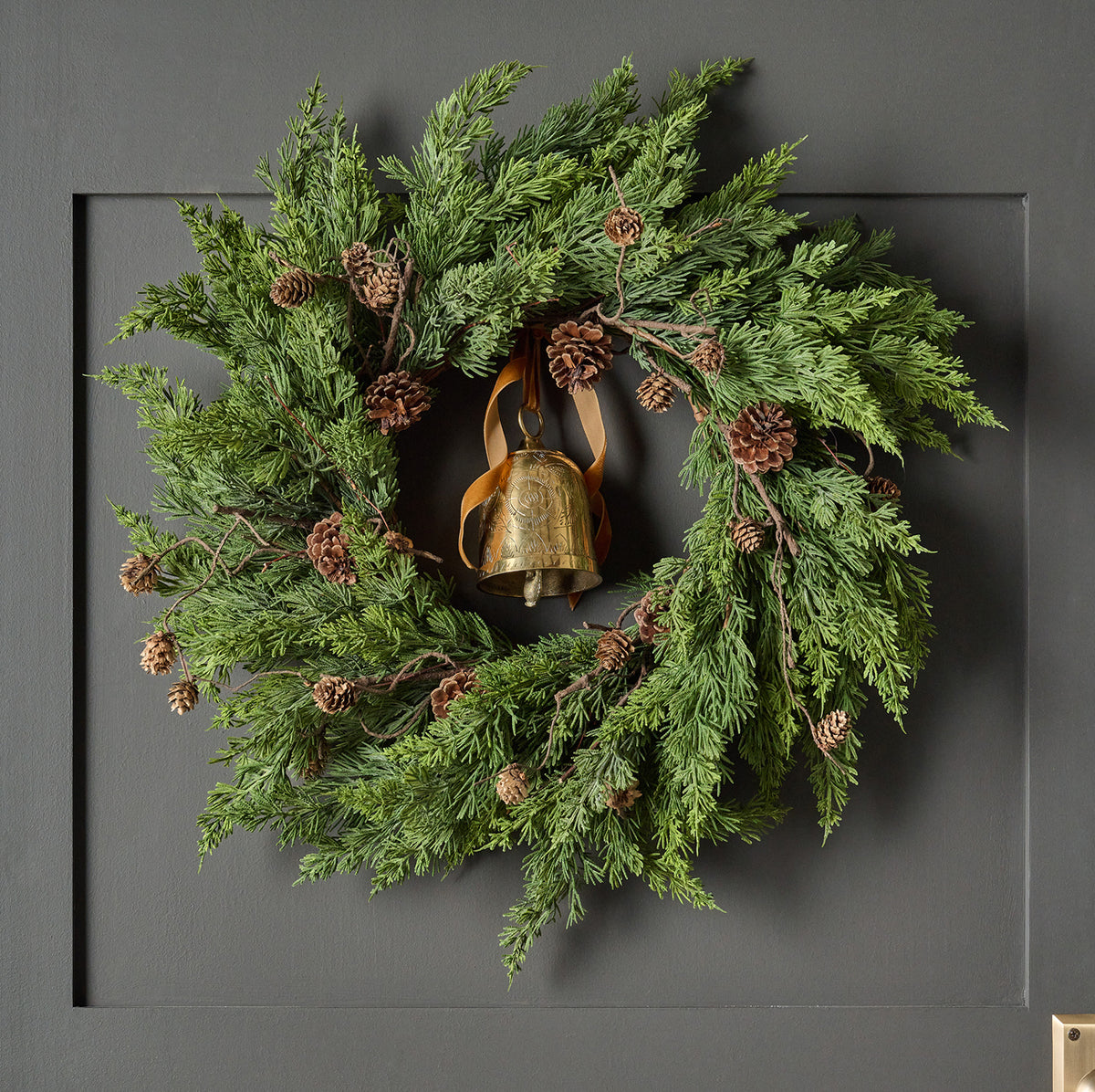 A green evergreen wreath with pinecones and a hanging brass bell, displayed on a dark gray paneled door.