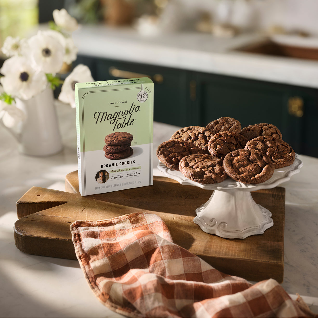 A box of Magnolia Table Frozen Brownie Cookies and a dessert plate full of baked brownie cookies next to it with a plaid red dishtowel nearby.