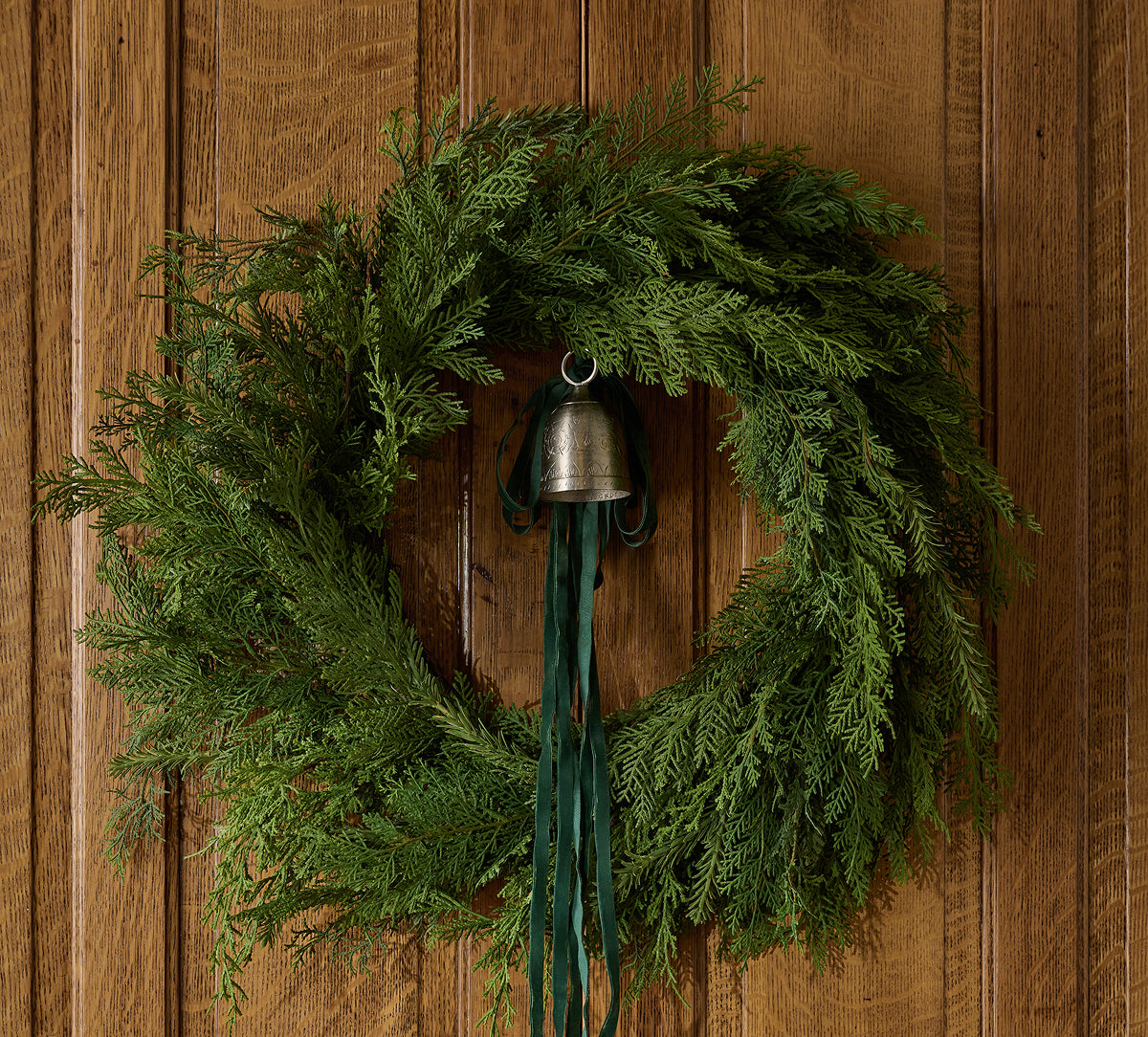 A green evergreen wreath hanging on a brown wooden door, with a small silver bell and dark green ribbon at the center.
