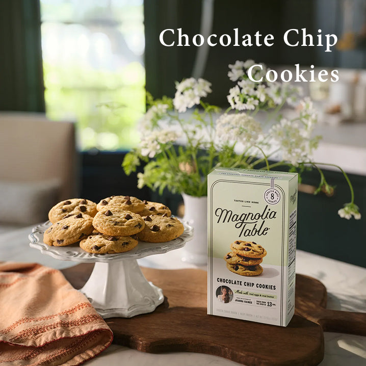Box of Magnolia Table Frozen Chocolate Chip Cookies on a kitchen island with baked chocolate chip cookies on a plate stand and flowers.  Chocolate Chip Cookies.  Learn More.