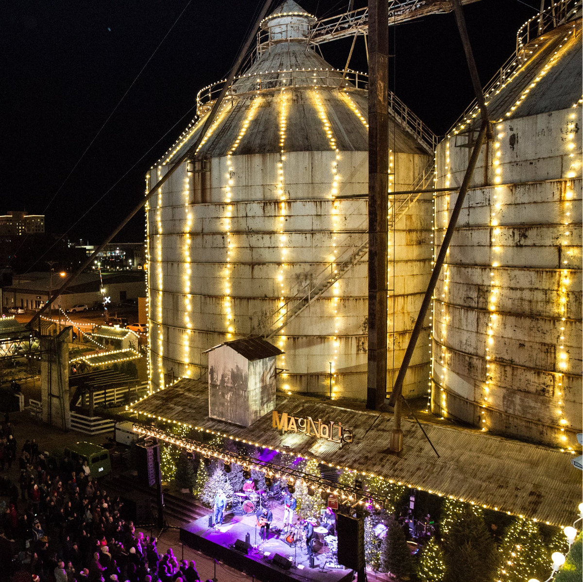 Nighttime aerial view of a large illuminated grain silo with warm yellow string lights, a small stage under a covered walkway, and a crowd gathered below in an outdoor market setting.