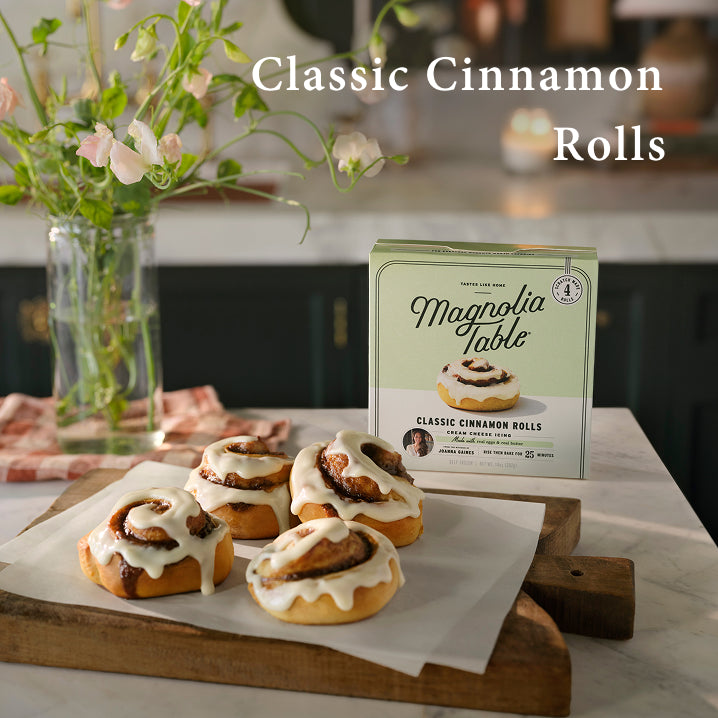 Box of Magnolia Table Frozen Classic Cinnamon Rolls on a kitchen island with a four baked classic cinnamon rolls on a cutting board and flowers. Classic Cinnamon Rolls.  Learn More.