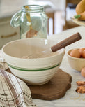 Beige mixing bowl with wooden handle on a kitchen counter with eggs and whisk.