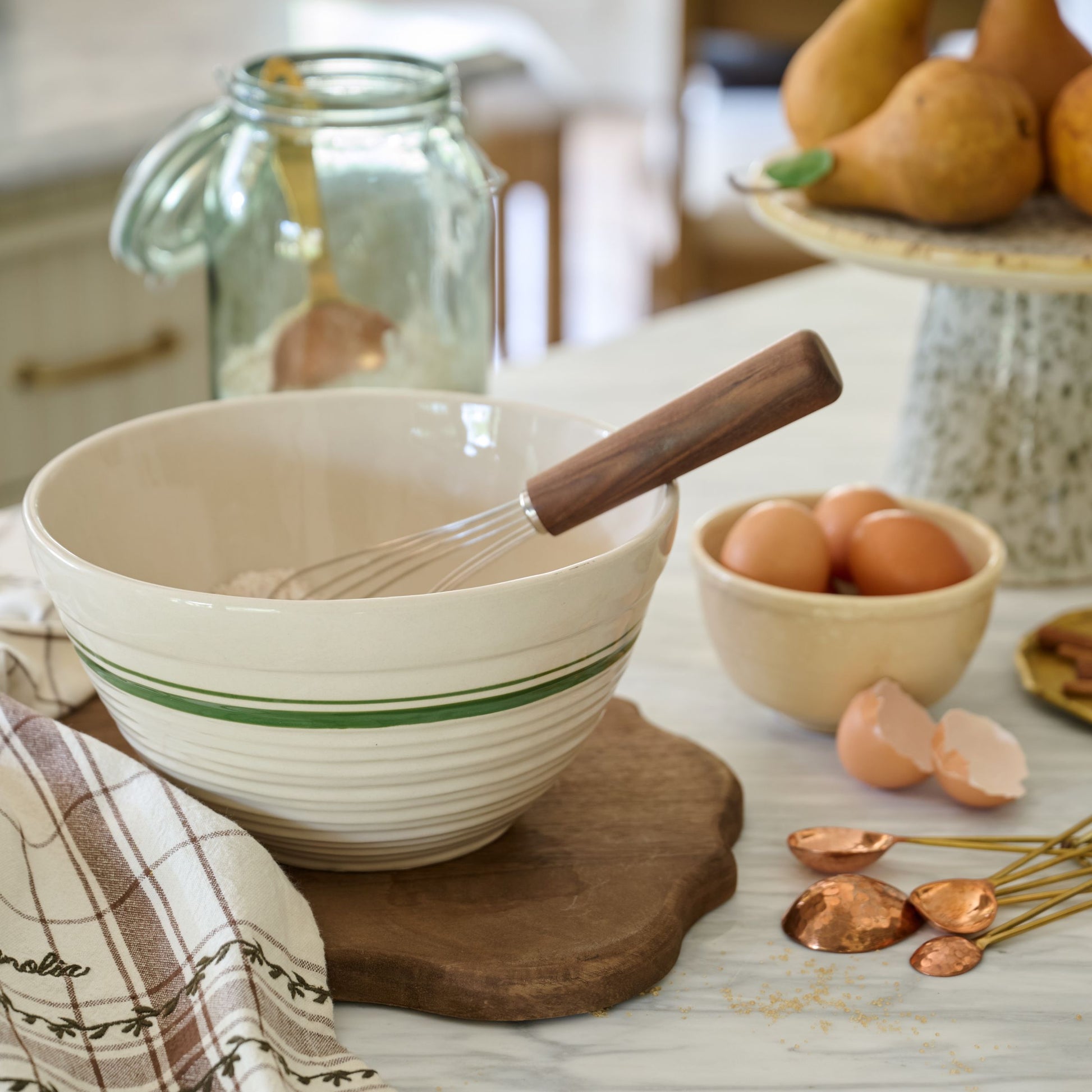 Beige mixing bowl with wooden handle on a kitchen counter with eggs and whisk.