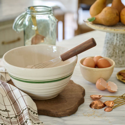 Beige mixing bowl with wooden handle on a kitchen counter with eggs and whisk.