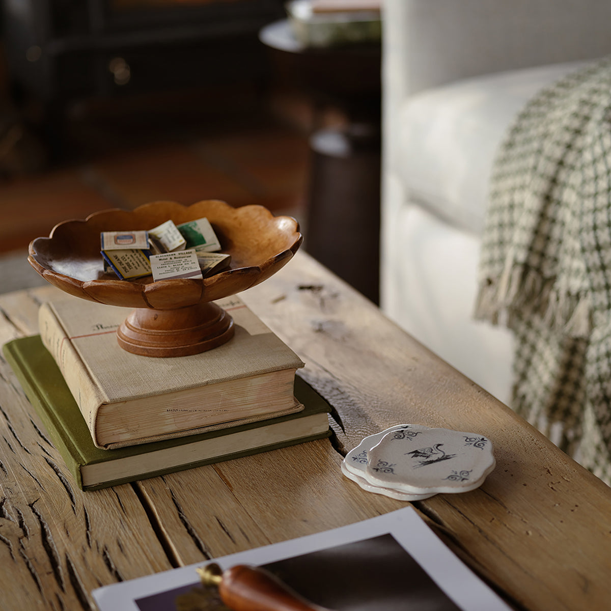 A scalloped wooden pedestal dish holding small vintage matchbooks sits on top of stacked books on a rustic wooden table, with a small white floral coaster nearby and a softly blurred living room background.