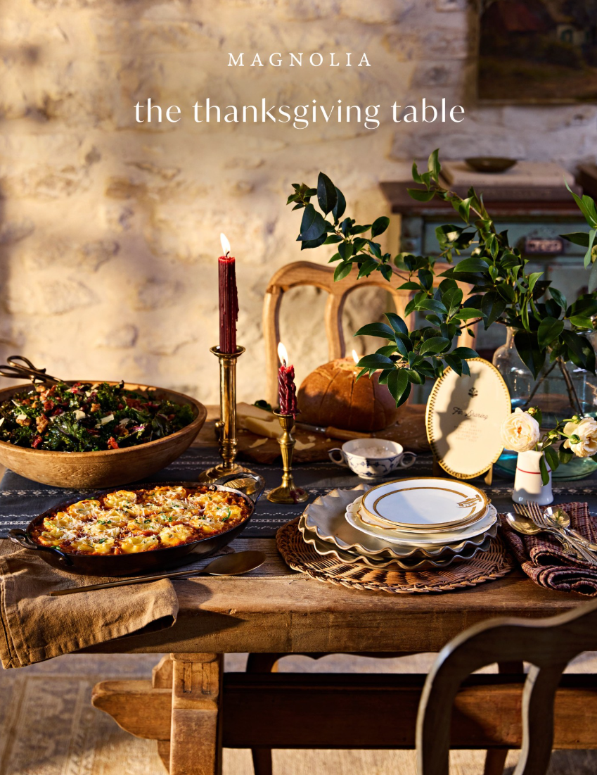 A styled Thanksgiving table scene with plated food, candles, plates, napkins, and greenery in a rustic indoor setting. The text 'MAGNOLIA' and 'the thanksgiving table' appears at the top.