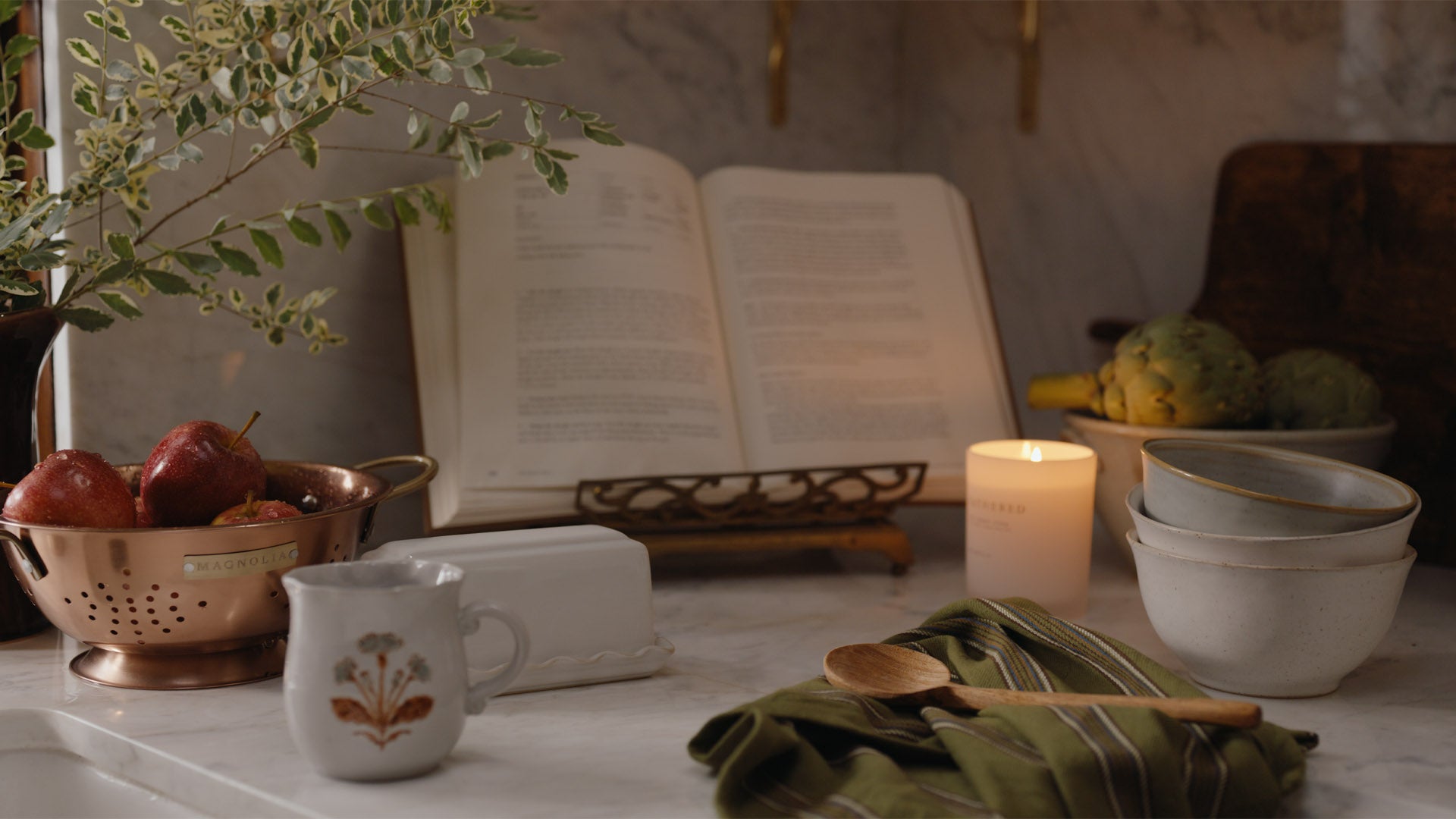 A styled tabletop scene with an open book on a stand, a lit candle, stacked ceramic bowls, a small white pitcher with a floral print, a green striped cloth, wooden spoons, and fruit in a copper colander, set against a softly blurred interior background.