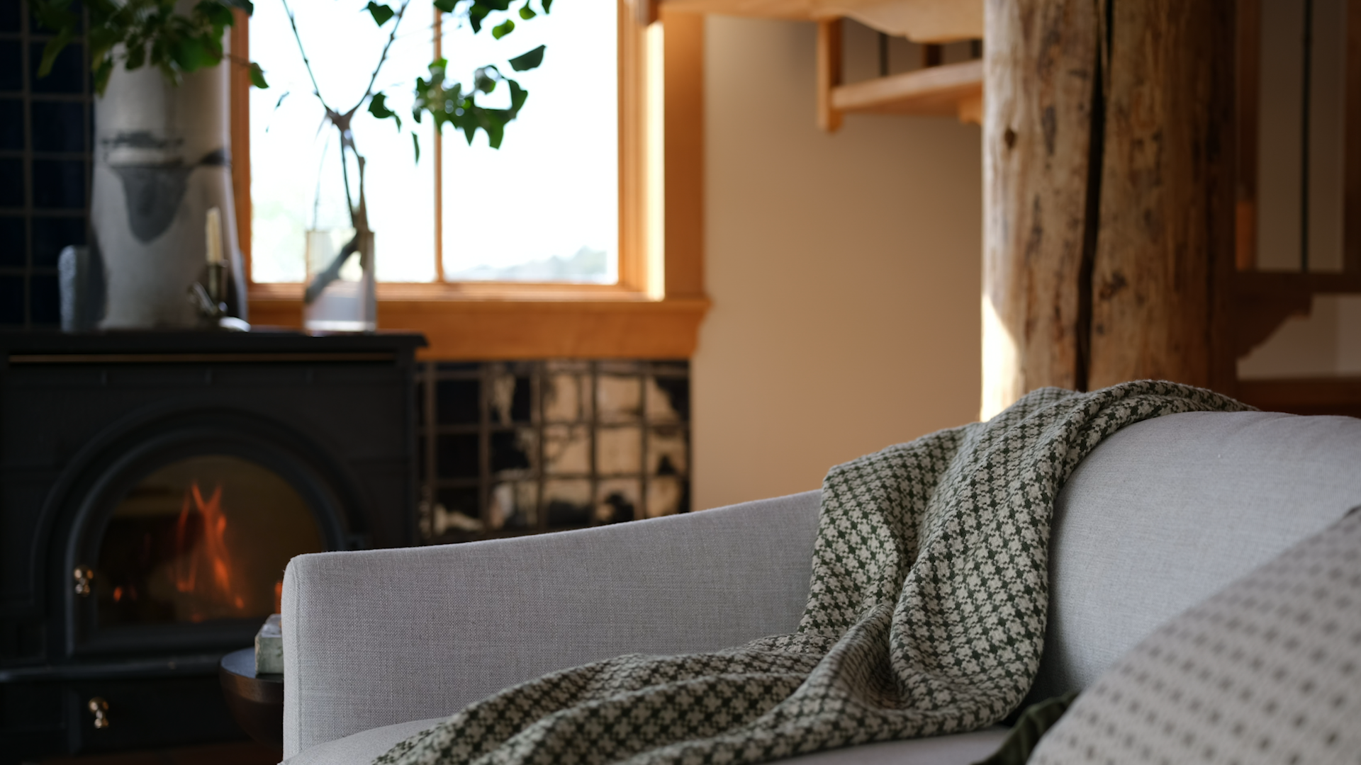 A close-up of a light gray upholstered armchair with a dark green and beige patterned throw blanket draped over it, set in a cozy room with a wood-burning fireplace, rustic wooden beams, and a window in the background.