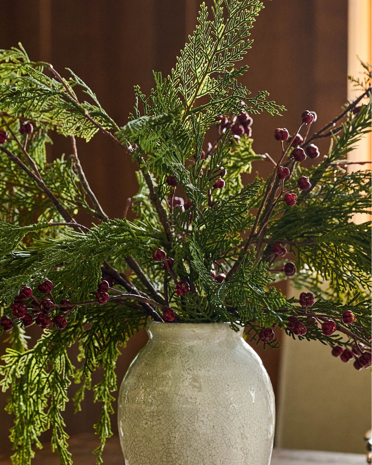 A rustic white ceramic vase filled with green evergreen branches and clusters of small dark red berries, set against a warm brown background.