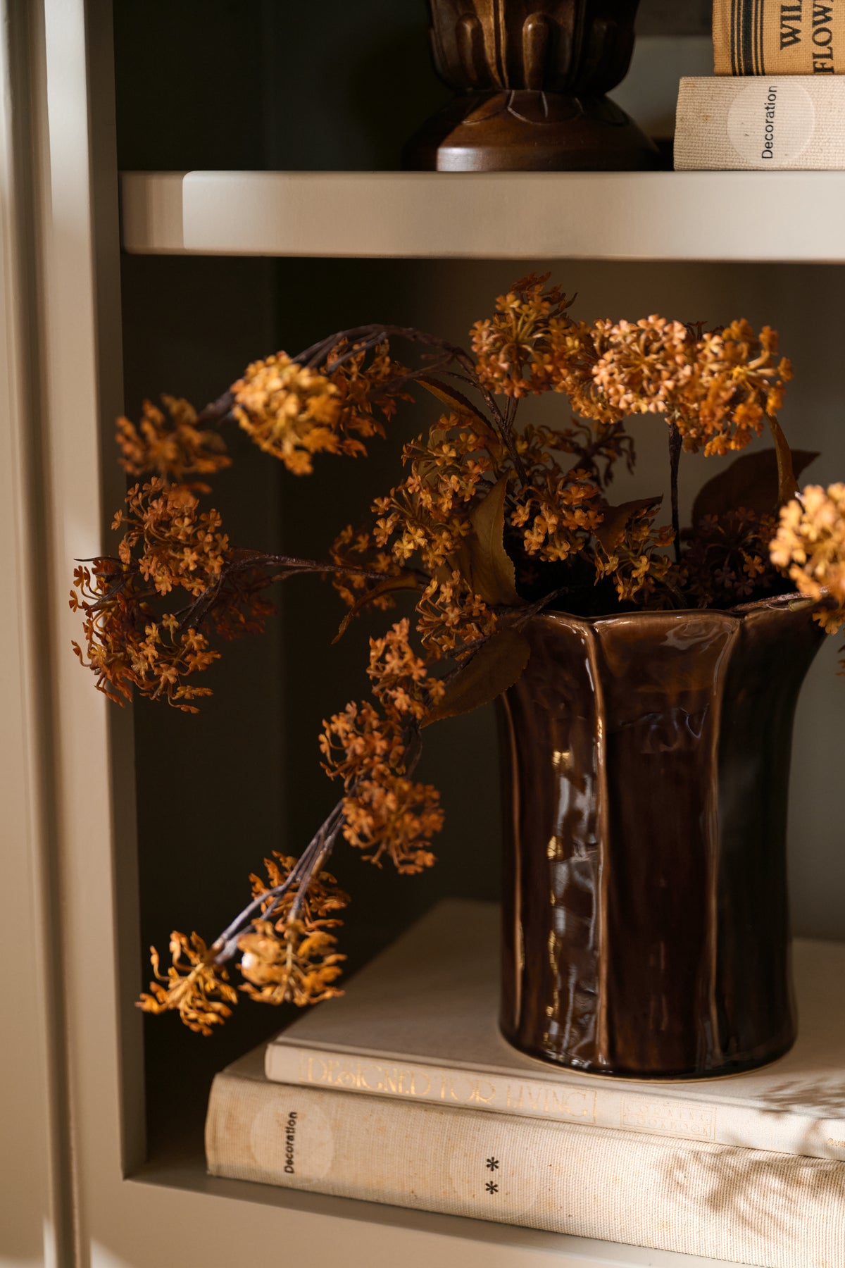 A dark brown glazed ceramic vase filled with dried orange-brown flowers, displayed on a bookshelf next to books.