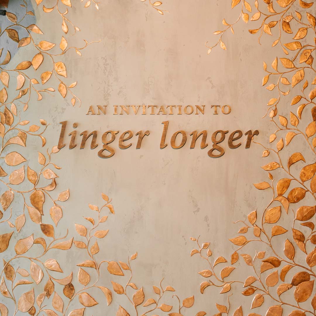 A square wedding invitation card design featuring the embossed text 'AN INVITATION TO linger longer' in a warm metallic bronze tone, surrounded by a border of metallic gold leafy vines on a textured beige background.
