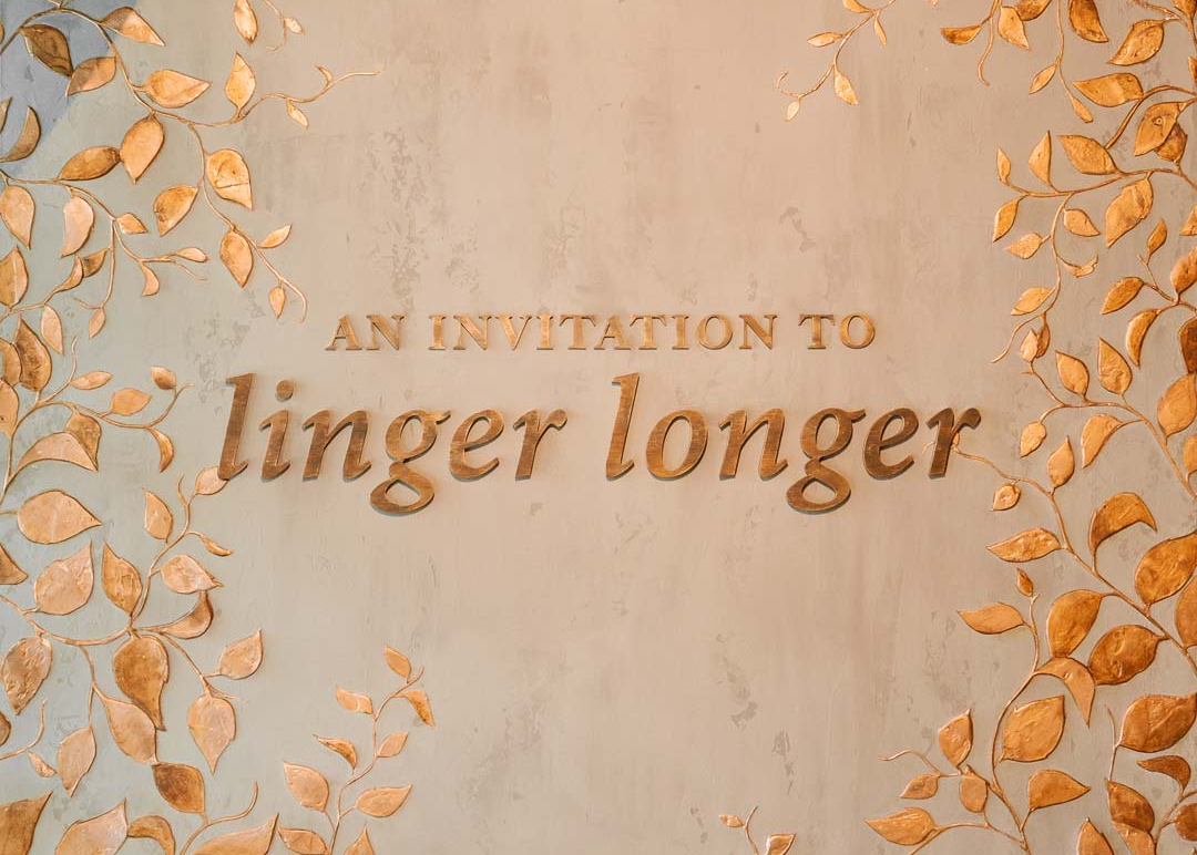 An elegant wedding invitation card design featuring the text 'AN INVITATION TO linger longer' in a warm metallic bronze font, surrounded by a border of 3D gold leaf vines on a textured beige background.