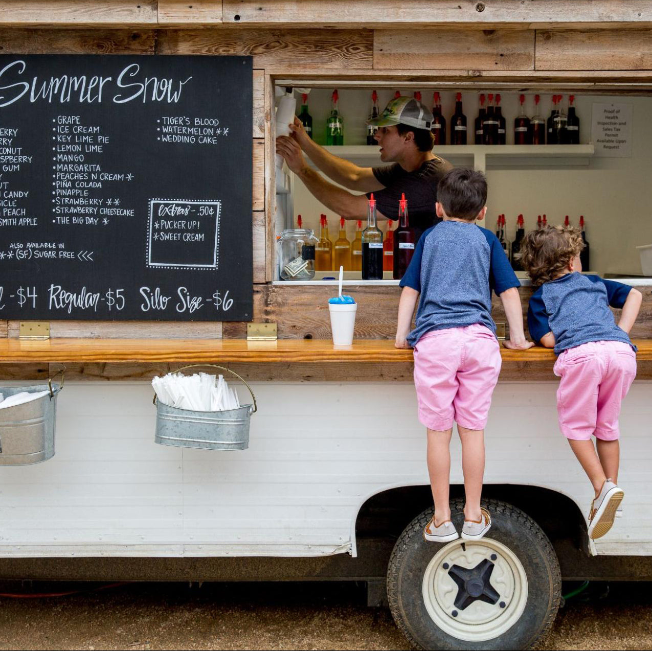 Two children standing on a food truck counter with a menu board in the background.