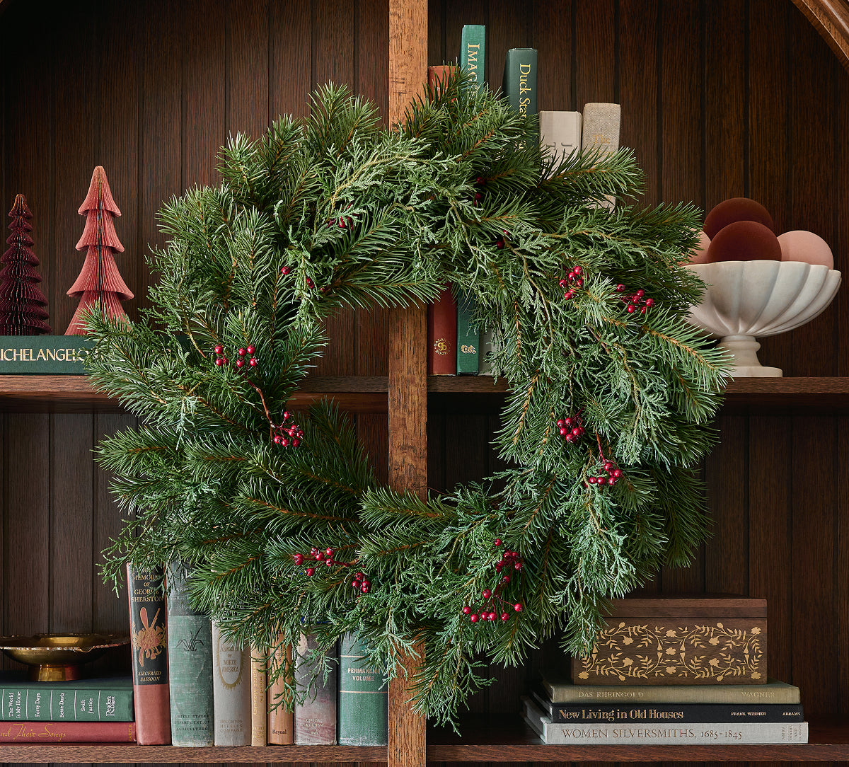 A green pine wreath with small red berry accents hanging on a wooden bookshelf, surrounded by books and holiday decor.