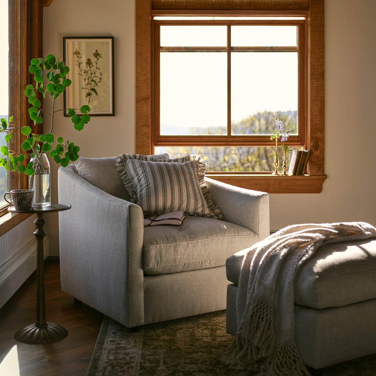 A light gray upholstered armchair in a sunlit living room, styled with striped and textured pillows, a side table with greenery, a window with wood trim, and a matching gray ottoman with a throw blanket.
