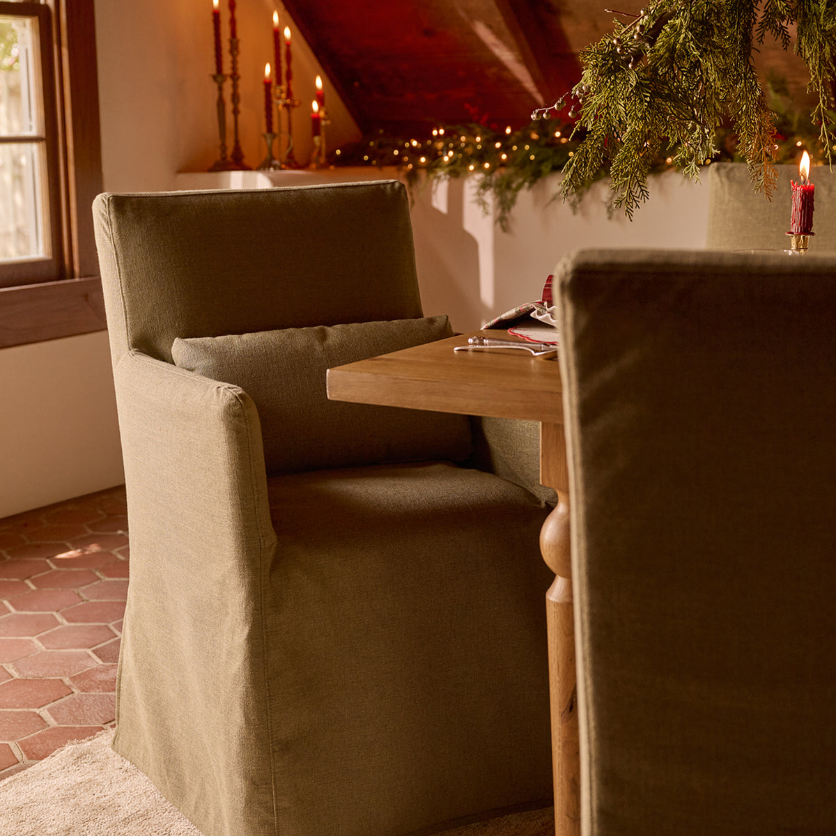 A rustic upholstered dining chair in a warm olive-brown fabric, styled in a cozy holiday home setting with a wooden table, candlelight, and festive greenery.