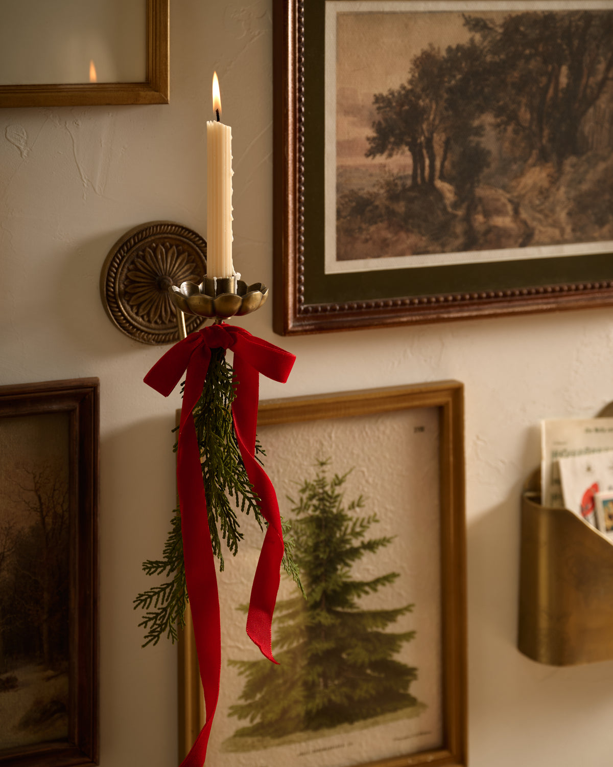 A lit white taper candle in a brass wall sconce, decorated with a red ribbon and green pine branches, mounted on a textured cream wall with framed landscape prints.