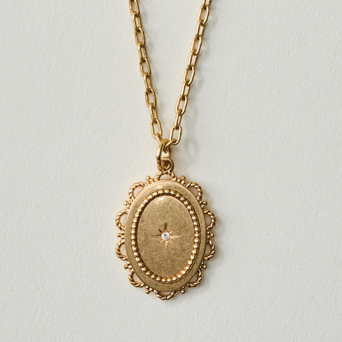 A gold oval locket pendant necklace with a delicate scalloped border and a small central clear stone, hanging against a light gray background.